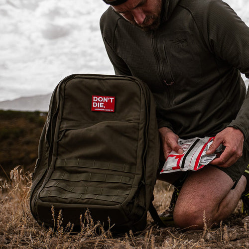 DON'T DIE Velcro Patch on olive green tactical backpack with MOLLE webbing, man holding survival food outdoors