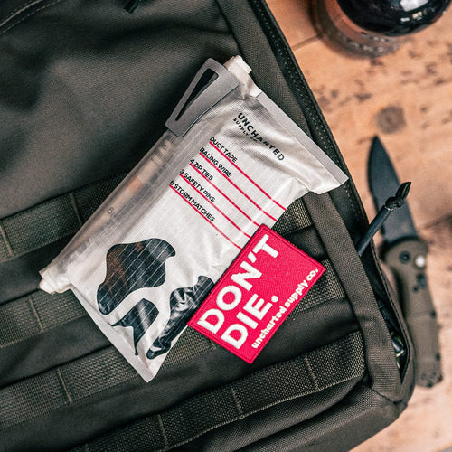 DON'T DIE Velcro Patch, red with white text, on an olive tactical backpack with a survival kit, essential survival gear