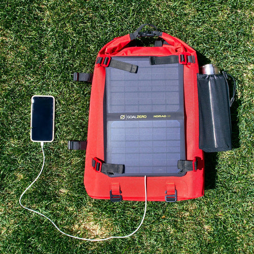 Goal Zero Nomad 10 Solar Panel charging a phone, attached to a red dry bag on grass, portable solar charger