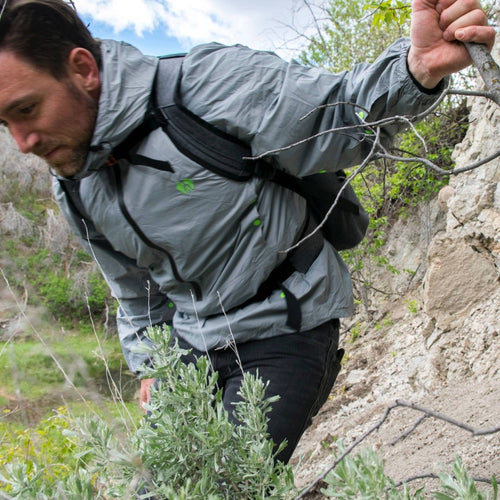 The Hideaway Weather-Resistant Windbreaker in gray on a man hiking uphill, gripping a branch for support