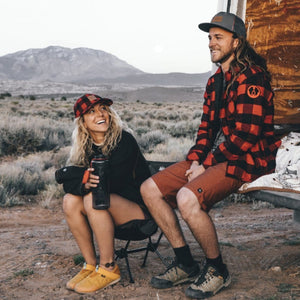 HWY8 Hat, a woman smiles wearing the red and black plaid cap in an outdoor camping scene with mountains