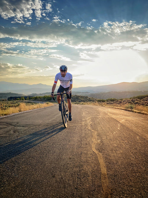A cyclist rides towards the camera wearing a white Uncharted Supply Co. cycling jersey.