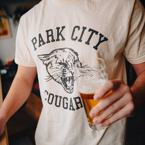 Park City Cougars T-shirt worn by a man holding beer, cream tee with black arched 